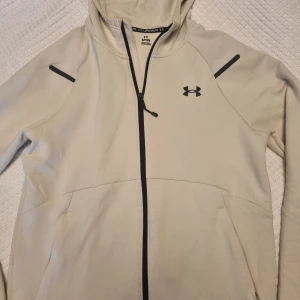 Ljusbeige hoodie från Under Armour - Ljusbeige hoodie från Under Armour med svart dragkedja och logga på bröstet. Jackan har huva och två fickor framtill, samt svarta detaljer på ärmarna. Inga byten. 