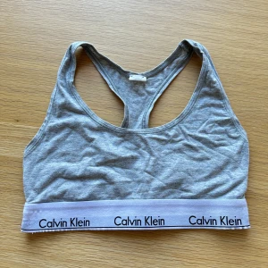 Grå sport-bh från Calvin Klein - Grå sport-bh från Calvin Klein med klassisk brottarrygg och vit resår med logga. Perfekt för träning eller som bekväm topp till vardags. Mjuk bomullsblandning och stilren design.