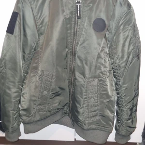 Grön bomberjacka med broderad text - Säljer en grön bomberjacka från Black squad med broderad text 'SEASON ONE' på ryggen. Jackan har dragkedja framtill, ribbade muddar och fickor med lock och knapp. Klassisk bomberstil med patchdetaljer.