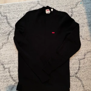 Svart stickad tröja från Levi's - Säljer en svart stickad tröja från Levi's med rund halsringning och diskret röd logga på bröstet. Tröjan har långa ärmar och ribbade muddar vid ärmslut och nederkant. Perfekt för lager-på-lager-stil.