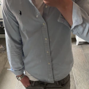 Ljusblå skjorta från Ralph Lauren - Säljer en klassisk ljusblå skjorta från Ralph Lauren med knappar framtill och broderad logga på bröstet. Skjortan har lång ärm och normal passform, perfekt till många olika tillfällen. Snygg och tidlös design!