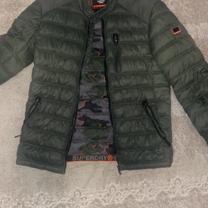 Grön pufferjacka från Superdry - Säljer en grön pufferjacka från Superdry med svart och orange logga på ärmen. Jackan har dragkedja framtill, två sidofickor med dragkedja och en bröstficka. Insidan har camouflagemönster med orange detaljer.