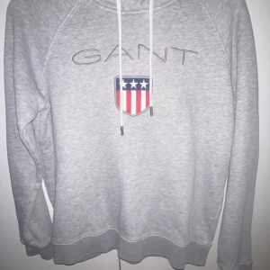 Grå hoodie från GANT med logga - Säljer en ljusgrå hoodie från GANT med broderad logga och sköld på bröstet. Tröjan har huva med dragsko och långa ärmar. Perfekt för en avslappnad och stilren look. Hör av er vid funderingar😁
