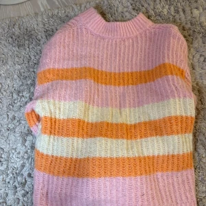Randig stickad tröja i rosa, orange och vit - Mysig stickad tröja med breda ränder i rosa, orange och vitt. Tröjan har ribbad struktur och hög hals, perfekt för kyligare dagar. Passar dig som gillar färg och vill ha en skön och trendig look.