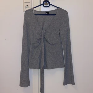 Grå långärmad topp med knyt från Gina Tricot - Säljer en grå långärmad topp från Gina Tricot med v-ringning och knyt framtill. Toppen har en figurnära passform och är tillverkad i mjukt bomullsmaterial. Perfekt för en stilren och enkel look. HELT NY, HAR ALDRIG ANVÄNTS 