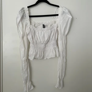 Vit blus med puffärmar från H&M - Säljer en vit blus med långa puffärmar och smockad midja. Toppen har fyrkantig ringning och är croppad med volangkant nedtill. Perfekt för en trendig och romantisk look.