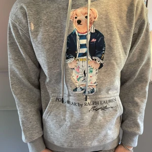 Grå Ralph Lauren hoodie - Tjena, Säljer nu en grå hoodie från Polo Ralph Lauren med ikoniskt björntryck på bröstet. Tröjan är i utmärkt skick och är perfekt för dig som vill ha en skön stilren tröja för ett dunder pris. 