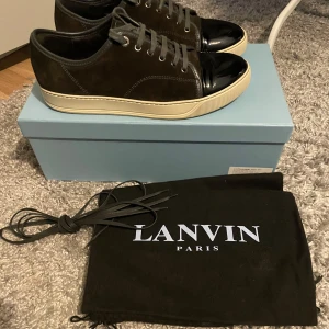 Svarta sneakers från Lanvin med lackad tå - Säljer ett par stilrena sneakers från Lanvin med snygg lackad tå och grå snörning. Skorna har en platt sula och kommer med extra skosnören samt originalpåse. Perfekta för dig som gillar exklusiva detaljer.