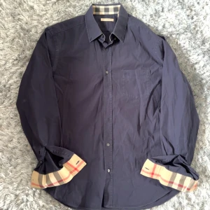 Mörkblå skjorta från Burberry med rutiga detaljer - Säljer en mörkblå skjorta från Burberry med klassiska beige-rutiga detaljer på insidan av krage och manschetter. Skjortan har en bröstficka och knäppning framtill. Perfekt för dig som gillar stilrena plagg med en twist.