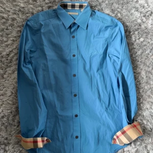 Blå skjorta från Burberry Brit - Snygg blå skjorta från Burberry Brit med klassiska rutiga detaljer på insidan av kragen och ärmsluten. Skjortan har knappar framtill och lång ärm. Perfekt för dig som vill ha en stilren look med en touch av lyx.