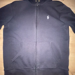 Svart hoodie från Polo Ralph Lauren - Säljer en svart hoodie från Polo Ralph Lauren med dragkedja och klassisk vit logga på bröstet. Tröjan har huva med snörning och två fickor framtill. Perfekt för en avslappnad stil.
