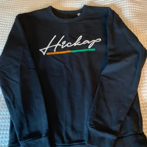Svart sweatshirt från Hickap - Svart sweatshirt från Hickap med tryckt logga i vitt, orange och grönt framtill. Klassisk rund halsringning och långa ärmar. Perfekt för en avslappnad stil.