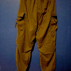 Bruna cargopants med stora fickor - Säljer ett par bruna cargopants med stora fickor på sidorna och dragsko i midjan. Byxorna har en loose passform och är perfekta för en avslappnad stil. Snygga detaljer med snörning vid fickorna och bensluten. Aldrig använda