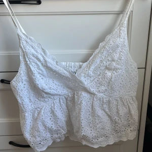 Vit broderad linnetopp från NA-KD - Säljer en söt vit linnetopp från NA-KD i storlek 38. Toppen har broderade blommor, spetsdetaljer och smala axelband. Perfekt till sommaren och har en lätt volang nertill samt smock i ryggen för skön passform.