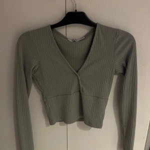 Ljusgrön ribbad croppad topp från NA-KD - Säljer en ljusgrön croppad topp från NA-KD med lång ärm och v-ringning. Toppen är ribbad och har en figurnära passform som framhäver siluetten. Perfekt att styla med jeans eller kjol.