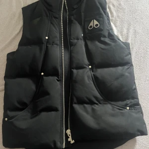 Svart dunväst från Moose Knuckles - Säljer en svart dunväst från Moose Knuckles med dragkedja framtill och två stora fickor. Västen har en quiltad design och logotyp på bröstet. Perfekt för lager-på-lager och håller dig varm under kyliga dagar. Endast använd 2-3 gånger 