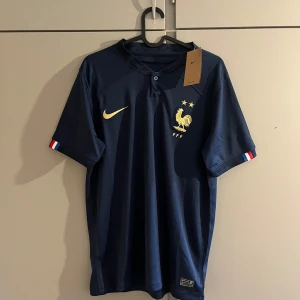 Frankrike fotbollströja Nike - Mörkblå officiell Frankrike fotbollströja från Nike med guldiga detaljer, inklusive Nike-logga och FFF-emblem med tupp och två stjärnor. Korta ärmar med franska flaggans färger på muddarna. Tillverkad i lätt och ventilerande material.