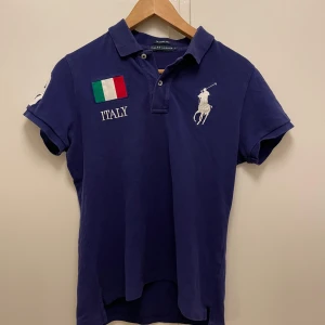 Blå pikétröja från Ralph Lauren med Italiens flagga - Säljer en blå pikétröja från Ralph Lauren i modellen The Skinny Polo. Tröjan har broderade detaljer med Italiens flagga och texten ITALY fram och bak, samt en stor vit siffra 3 på ärmen och ryggen. Klassisk krage och korta ärmar.