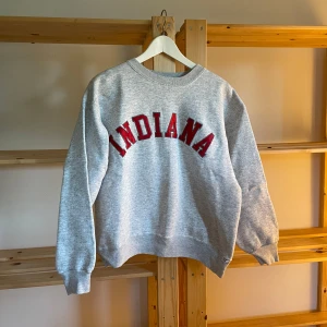 Grå sweatshirt med Indiana-tryck - Säljer en grå sweatshirt med rött 'INDIANA'-tryck över bröstet. Klassisk rund halsringning 