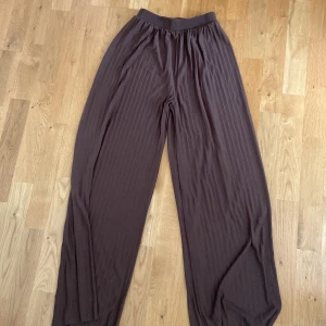 Bruna vida byxor med resårmidja - Säljer ett par bruna vida byxor med resår i midjan. Byxorna har ett diskret randigt mönster och är tillverkade i ett mjukt, lätt material som ger en skön känsla. Perfekta för en avslappnad och trendig stil. Ganska stora i storlek 