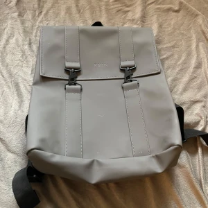 Grå ryggsäck från Rains - Snygg och stilren grå ryggsäck från Rains med två spännen framtill och justerbara svarta axelband. Perfekt för vardagsbruk eller när du behöver bära med dig lite extra. Vattentåligt material och minimalistisk design.