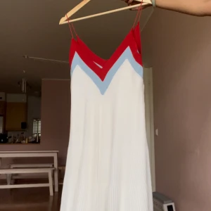 Vit plisserad klänning med röd och ljusblå detalj - Säljer en vit plisserad långklänning med smala axelband och snygga v-formade detaljer i rött och ljusblått upptill. Klänningen har en flowy känsla och är perfekt för sommarens tillfällen. Ingen synlig logga eller märke.