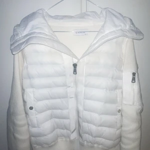 Vit pufferjacka från Moncler med huva - Säljer en snygg vit pufferjacka från Moncler med stickade ärmar och huva. Jackan har dragkedja framtill, två fickor med knappar och en ärmficka med dragkedja. Klassisk Moncler-logga på ärmen och serietidningsdetalj på insidan, NFC Scan funkar, Storlek S passar M.