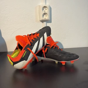Adidas Predator fotbollsskor svart/vit/röd - Säljer ett par Adidas Predator fotbollsskor i svart med vita ränder och röda detaljer. Skorna har snörning, låg profil och platt sula med dobbar för bra grepp på planen. Perfekta för dig som vill ha kontroll och stil på fotbollsplanen. Skorna är i toppskick och har inga defekter, krävs endast en skön tvätt så är dom som nya!!!