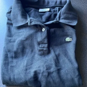 Svart pikétröja från Lacoste - Klassisk svart pikétröja från Lacoste med broderad krokodillogga på bröstet. Tröjan har korta ärmar, krage och knappar framtill. Perfekt för en stilren och sportig look.