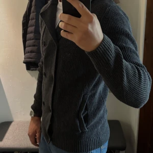 Mörkgrå stickad kofta med knappar - Mörkgrå stickad kofta med dubbelknäppning och bred krage. Koftan har långa ärmar och två fickor framtill. Perfekt för lager-på-lager och passar till jeans eller chinos.