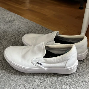Vita slip-on sneakers från Vans - Säljer ett par klassiska vita slip-on sneakers från Vans. Skorna har en enkel och stilren design utan snörning, med rund tå och platt sula. Perfekta för dig som gillar en clean och avslappnad stil.