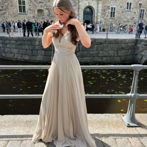 Champagnefärgad långklänning med pärldetaljer - Elegant långklänning i champagnefärg med glittrig, pärlbroderad överdel och djup v-ringning. Klänningen har snörning i ryggen samt en liten dragkedja för bättre passform. Perfekt för bal eller fest.