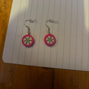Örhängen med drakfrukt-design - Säljer ett par unika örhängen som ser ut som skivor av drakfrukt. De är runda med vit mitt, svarta prickar och en stark rosa kant. Perfekta för dig som vill ha något färgglatt och lekfullt i öronen!