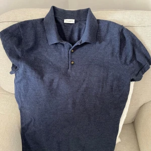 Mörkblå pikétröja i merinoull från John Henric - Säljer en stilren mörkblå pikétröja från John Henric i 100% merinoull. Tröjan har klassisk krage, två knappar framtill och korta ärmar. Perfekt för dig som vill ha en bekväm och snygg tröja med exklusiv känsla.