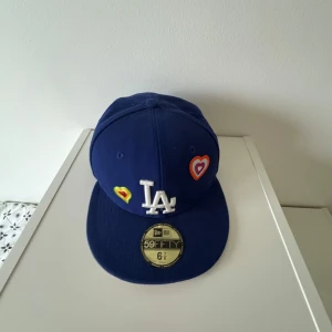 Blå keps från New Era 59FIFTY LA med broderade hjärtan - Storlek: 54.9 cm   Har använd kepsen men har inga skador. Men har färgats av lite på den rosa färgen men det går säkert bort efter man har tvättat den.  Har hjärtan med olika färger och klistermärket är det originella. Köpte kepsen på Hatstore för 550kr