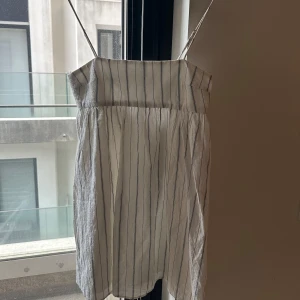 Vit och svart randig sommarklänning med tunna axelband - Säljer en söt vit klänning med svarta ränder och tunna axelband. Klänningen har en rak överdel och faller löst från bysten, perfekt för varma dagar. Enkel och stilren design som passar till många tillfällen.