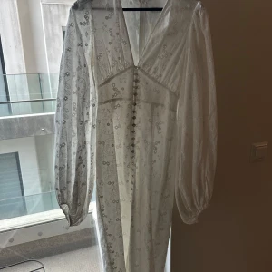 Vit långklänning med spetsdetaljer och knappar - Romantisk vit långklänning med v-ringning, långa puffärmar och dekorativa knappar framtill. Klänningen har fina spetsdetaljer och ett diskret blommigt mönster. Perfekt för sommarens speciella tillfällen.