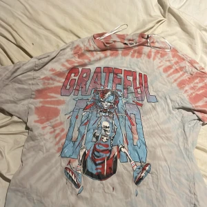 Grateful Dead tie dye t-shirt - Säljer en vintageinspirerad tie dye t-shirt med Grateful Dead-tryck framtill. T-shirten har röd och ljusblå spiralbatik, korta ärmar och en stor färgglad grafik med skelett och texten 'GRATEFUL'. Perfekt för dig som gillar unik och retro stil.