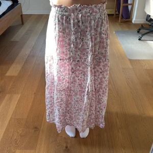 Blommig långkjol från AWARE - Säljer en söt långkjol från AWARE med blommigt mönster i rosa och vitt. Kjolen har resår i midjan och är luftig med ett flowy fall, perfekt för varmare dagar. Superfin att styla med en enkel topp eller stickad tröja.