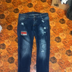 Mörkblå jeansbyxor från Dsquared2 - Säljer ett par mörkblå jeansbyxor från Dsquared2 med slitningar, färgstänk och en röd patch på ena benet. Klassisk femficksmodell med snygga detaljer och Dsquared2-logga bak i midjan. Perfekta för dig som vill sticka ut lite extra.