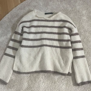 Vit och brunrandig stickad tröja från Vero Moda - Mysig stickad tröja från Vero Moda i vitt med bruna horisontella ränder. Tröjan har lång ärm och en rak, något boxig passform. Perfekt för lager-på-lager och enkel att matcha med jeans eller kjol.