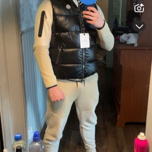 Svart dunväst från Moncler - Snygg svart dunväst från Moncler med glansig yta och dragkedja framtill. Perfekt för att hålla värmen med stil. Västen har Monclers ikoniska logga på bröstet.