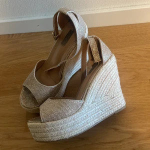 Glittriga platåsandaler med kilklack - Säljer ett par snygga sandaler med hög kilklack och platåsula i repimitation. Skorna är glittriga i en ljusbeige nyans och har öppen tå samt justerbart spänne runt vristen. Perfekta för dig som vill sticka ut lite extra!