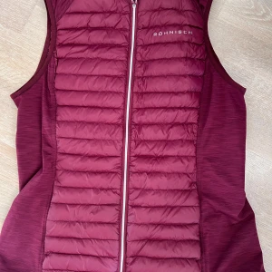 Vinröd dunväst från Röhnisch - Snygg vinröd dunväst från Röhnisch med quiltad framsida och baksida. Västen har dragkedja framtill och en sportig look, perfekt för lager-på-lager. Storlek M.