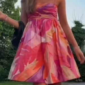 Färgglad kortklänning med abstrakt mönster - Säljer en färgglad kortklänning med abstrakt mönster i rosa, orange, lila och rött. Klänningen har smala axelband, markerad midja och utställd kjol. Perfekt för varma dagar och somriga tillfällen.