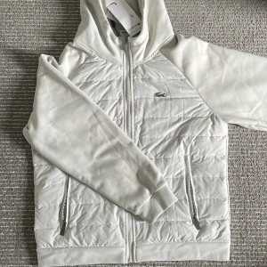 Vit cardigan från Lacoste - Snygg vit hoodie från Lacoste med quiltad framsida och dragkedja. Jackan har långa ärmar, två fickor med dragkedja och klassisk krokodillogga på bröstet. Perfekt för en stilren och sportig look. Passar även dig som har M.