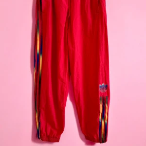 Röda Adidas Originals mjukisbyxor med regnbågsränder / storlek M /  -  🍓Sporty meets trendy – a must-have for 2025 street style! 🎀 Coquette meets athleisure – style with a baby tee & ballet flats. 🏃‍♀️ Super comfy – elastic waist with drawstring for a perfect fit. 🖤 Classic Adidas vibes – iconic side stripes & ribbed cuffs. ✨ Preloved in great condition – ready for a new wardrobe! 📏 Waist ~ 35 cm 📏 Leg length ~ 70 cm (internal) // 102 cm (external)