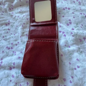 Vinrött sminkfodral med dekorativ knapp - Säljer ett vinrött sminkfodral i skinn med vit kontrastsöm och en snygg dekorativ blomformad metallknapp framtill. Fodralet har en sminkspegel inuti & är en perfekt accessoar för dig som vill ha något stilrent och unikt.
