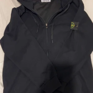 Svart hoodiejacka från Stone Island - Svart hoodiejacka från Stone Island med dragkedja framtill och klassisk logotyp på ärmen. Jackan har huva med dragsko och ribbade muddar vid ärmslut och nederkant. Perfekt för en avslappnad och stilren look. Pris kan diskuteras vid snabb affär
