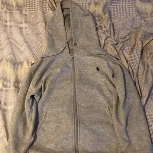 Grå zip hoodie från Polo Ralph Lauren - Säljer en klassisk grå hoodie från Polo Ralph Lauren med dragkedja och huva. Tröjan har långa ärmar, två fickor fram och den ikoniska lilla broderade loggan på bröstet. Perfekt för en avslappnad stil.
